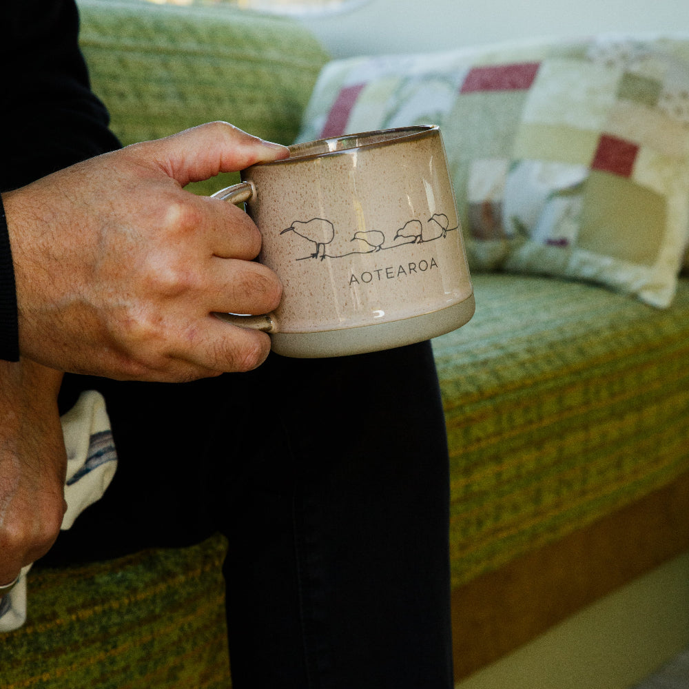 Glazed Ceramic Mug Kiwi Brown . Person holding a ceramic mug with 'Aotearoa' text and sheep design in a home setting.