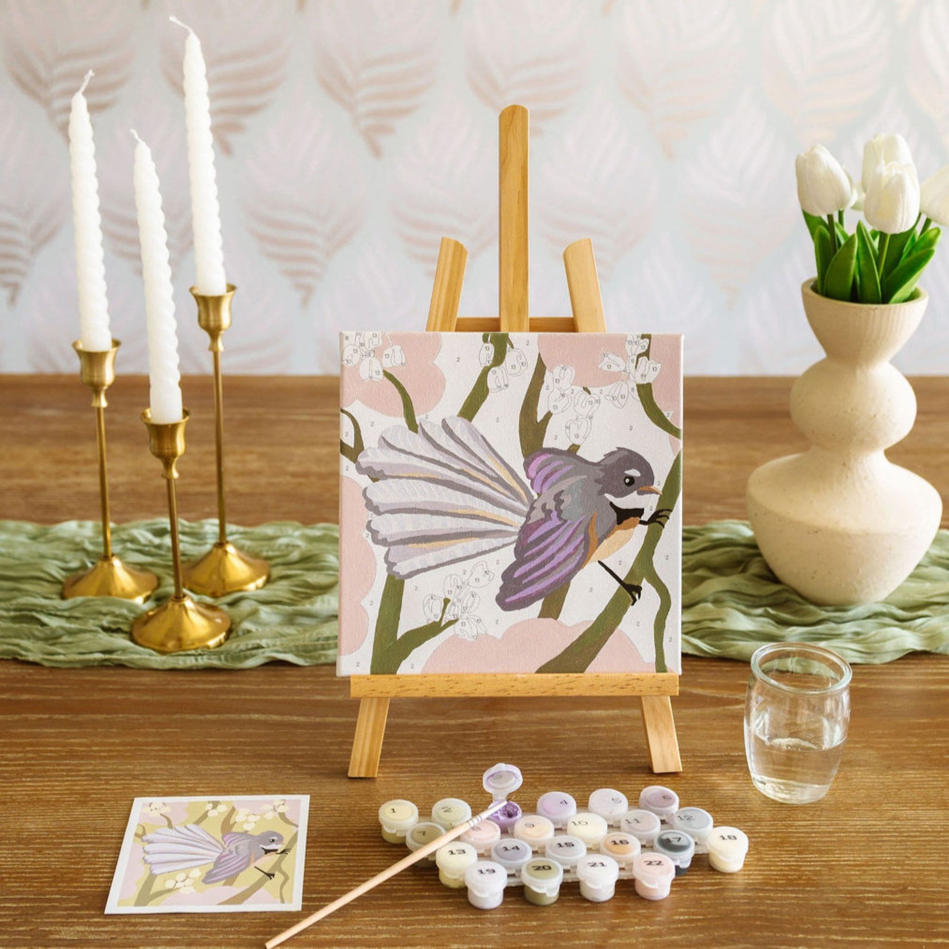 Artistic setup with a painting of a bird on an easel, candles, and art supplies on a wooden surface.
Adventure Collections  Fantail Paint By Numbers Kit – 20 x 20cm