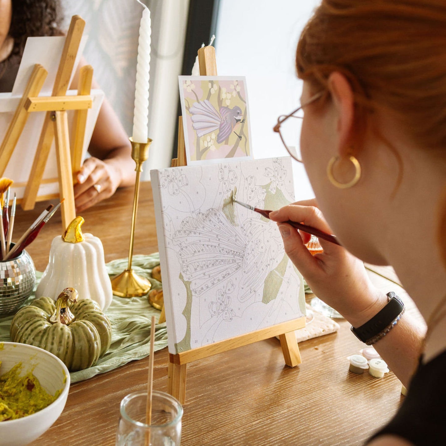 Person painting a small canvas with a bird design on a wooden table. Adventure Collections  Fantail Paint By Numbers Kit – 20 x 20cm