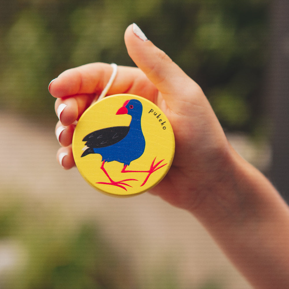 Hand holding a circular yellow yoyo with a colorful pukeko bird design against a blurred green background