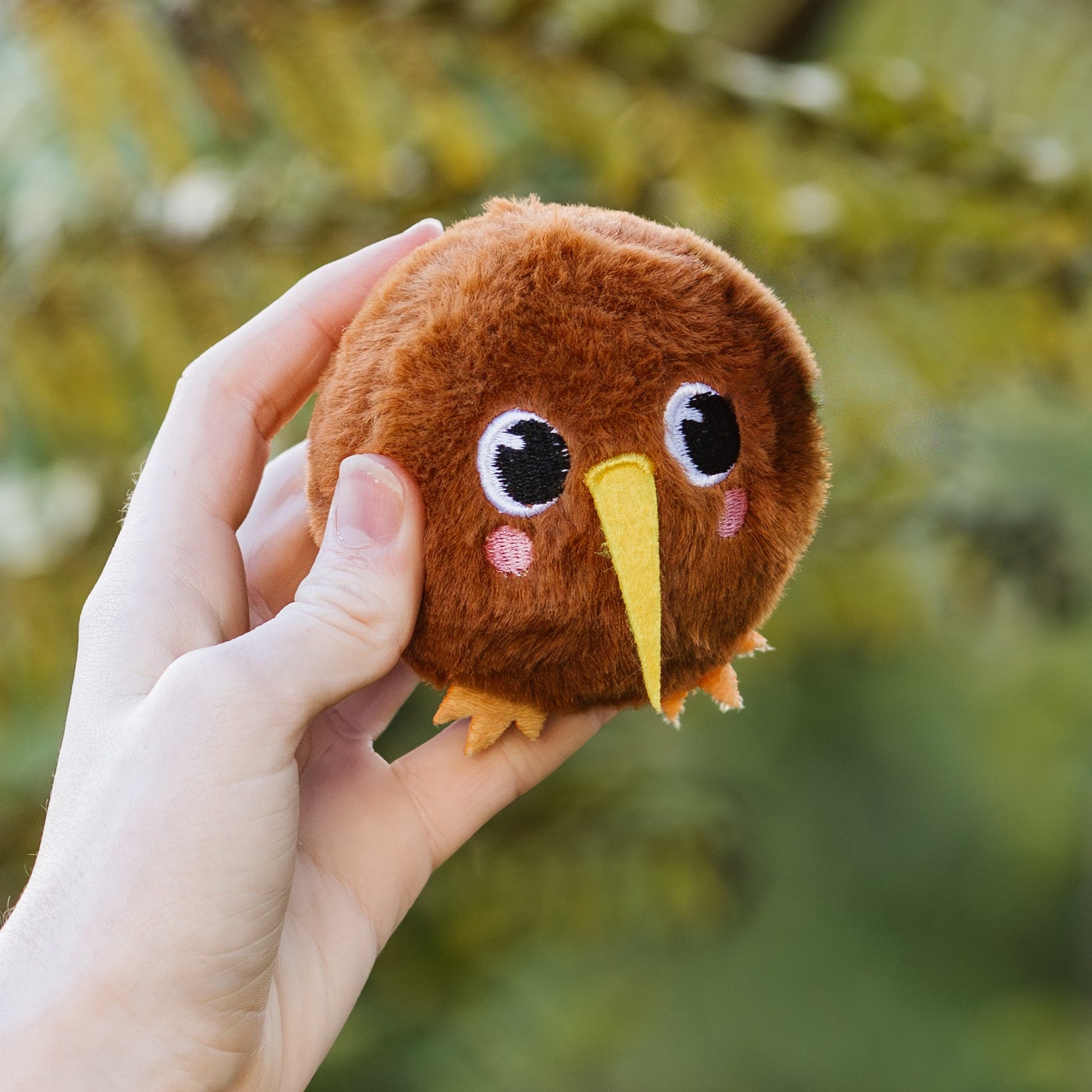Plush kiwi bouncy ball toy held in a hand with a blurred green background