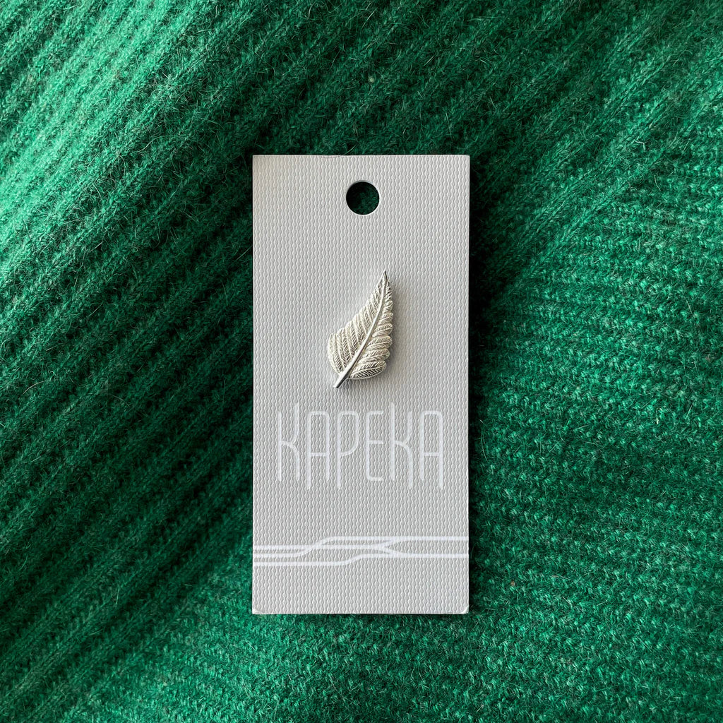 Silver leaf-shaped pin on a gray card against a green textured background.