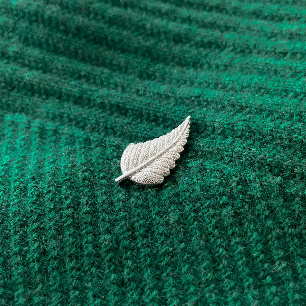 A silver pin shaped like a fern leaf resting on a green textured surface.