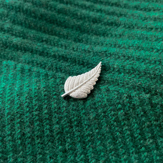 A silver pin shaped like a fern leaf resting on a green textured surface.