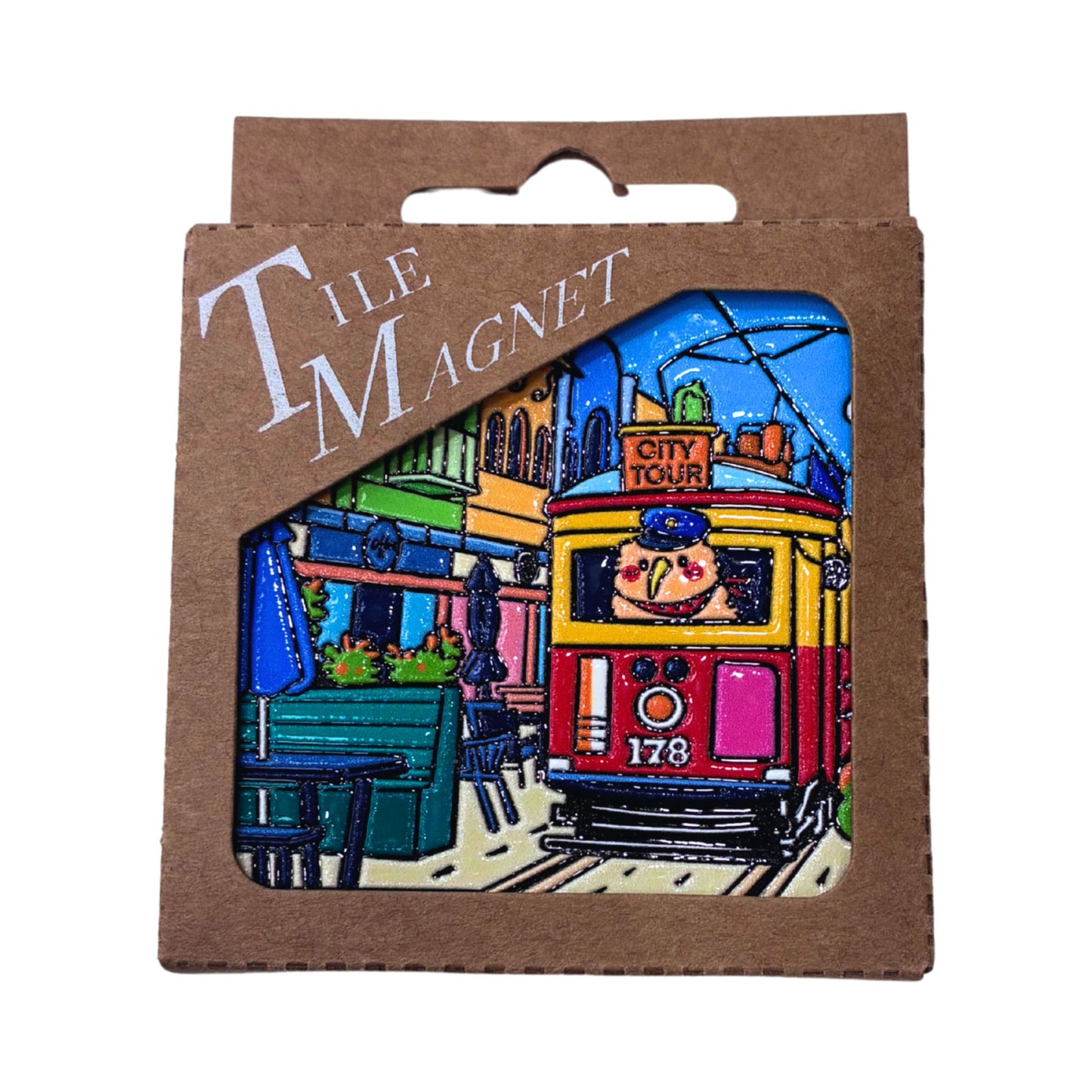 A colorful illustrated tile magnet in its brown cardboard retail packaging, featuring a vintage tram scene with the words 'TILE MAGNET' and 'CITY TOUR'.