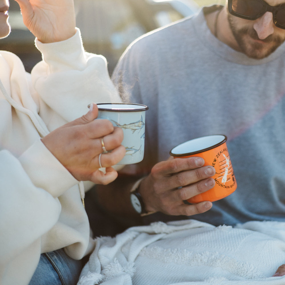 Two people sitting outdoors, holding moana road enamel mugs, wrapped in a blanket.