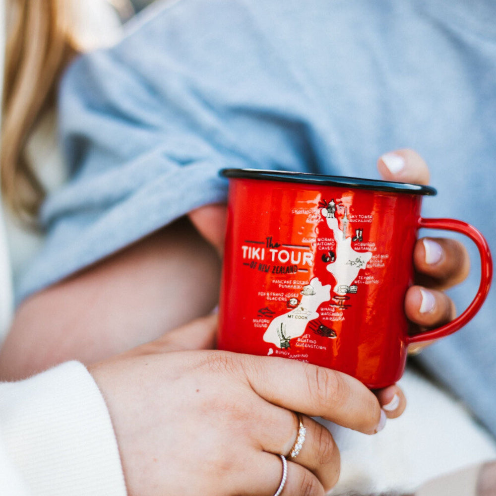Red tiki tour enamel mug with a map design held by two hands, one wearing a ring.